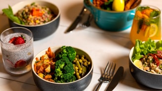 A table is set with several dishes containing a variety of healthy foods. There are bowls filled with colorful salads, vegetables like broccoli and peas, grains topped with vegetables, and glasses of infused water garnished with strawberry slices. A fork and knife are placed on the table, suggesting a meal ready to be enjoyed.