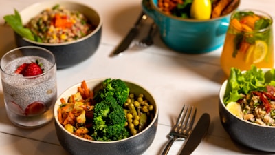 A table is set with several dishes containing a variety of healthy foods. There are bowls filled with colorful salads, vegetables like broccoli and peas, grains topped with vegetables, and glasses of infused water garnished with strawberry slices. A fork and knife are placed on the table, suggesting a meal ready to be enjoyed.