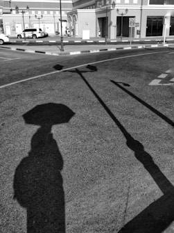 Shadows of street signs and a person with an umbrella fall on an asphalt surface. The background includes a street with parked vehicles and buildings with arched entrances and multiple windows.