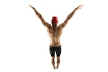 A muscular individual is viewed from behind, standing with arms extended upward forming a V shape. The person is wearing a red swimming cap and dark swimming shorts, set against a plain white background.