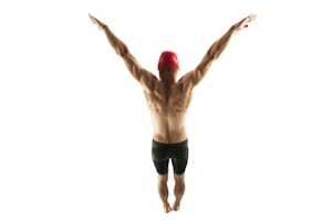 A muscular individual is viewed from behind, standing with arms extended upward forming a V shape. The person is wearing a red swimming cap and dark swimming shorts, set against a plain white background.