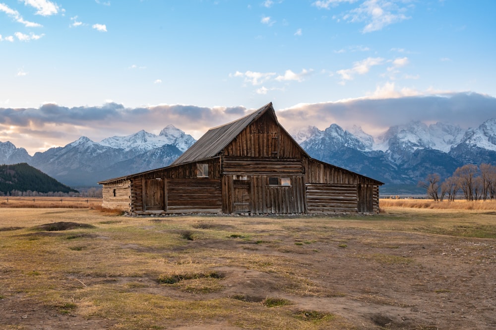 昼間は青空の下、雪に覆われた山々の近くの茶色の野原にある茶色の木造納屋