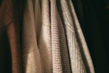 A row of textured clothing items hangs closely together. The fabrics vary in weave and color, with patterns ranging from smooth to coarse.