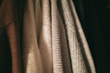 A row of textured clothing items hangs closely together. The fabrics vary in weave and color, with patterns ranging from smooth to coarse.
