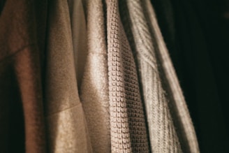 A row of textured clothing items hangs closely together. The fabrics vary in weave and color, with patterns ranging from smooth to coarse.