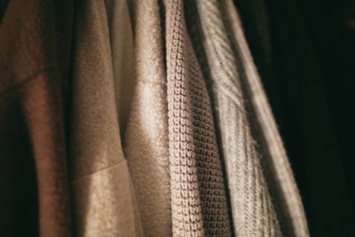 A row of textured clothing items hangs closely together. The fabrics vary in weave and color, with patterns ranging from smooth to coarse.
