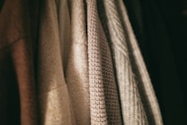 A row of textured clothing items hangs closely together. The fabrics vary in weave and color, with patterns ranging from smooth to coarse.