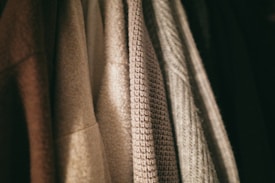 A row of textured clothing items hangs closely together. The fabrics vary in weave and color, with patterns ranging from smooth to coarse.