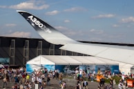 A large airplane wing with the label 'A350 XWB' is prominently visible in an outdoor exhibition area. Below the wing, a large gathering of people can be seen walking and socializing around a display area with tents. The area is cordoned off with barriers, and there are several informational booths or displays. The sky above is mostly clear with a few clouds.