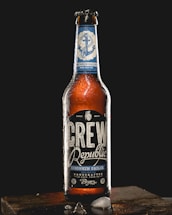 A bottle of Crew Republic Drunken Sailor India Pale Ale stands upright on a wooden surface against a dark background. The bottle is covered with condensation, and the label features nautical design elements including an anchor. Pieces of ice are scattered on the wooden surface.