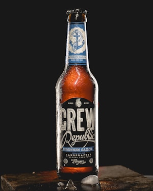 A bottle of Crew Republic Drunken Sailor India Pale Ale stands upright on a wooden surface against a dark background. The bottle is covered with condensation, and the label features nautical design elements including an anchor. Pieces of ice are scattered on the wooden surface.