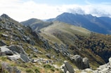 A group of hikers is walking along a rugged mountain trail, surrounded by rocky outcrops and green vegetation. The landscape features towering mountain peaks and a dense forested area on one side. The sky is partially cloudy, casting soft shadows over the terrain.