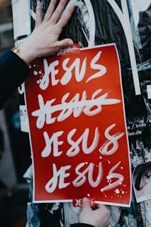 A red poster with the word 'Jesus' written in bold white letters multiple times. Several of the words are crossed out with red lines. A person is holding the poster with their hands, one of which is wearing a watch. The poster is placed on a surface covered with other printed papers and torn posters.