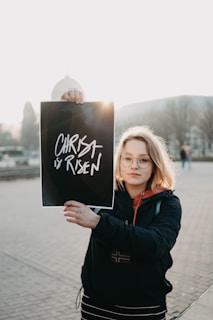 A person with shoulder-length blonde hair and glasses holds up a sign with the words 'Christ is Risen' written on it. They are standing in an outdoor setting with a paved area and blurred background of buildings and trees. The individual is wearing a black jacket and appears to be in a public space.