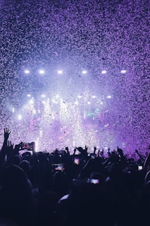 A vibrant concert scene with a large crowd raising their hands and holding up phones. The air is filled with confetti illuminated by colorful stage lights, creating a festive atmosphere.
