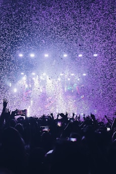 A vibrant concert scene with a large crowd raising their hands and holding up phones. The air is filled with confetti illuminated by colorful stage lights, creating a festive atmosphere.