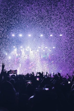 A vibrant concert scene with a large crowd raising their hands and holding up phones. The air is filled with confetti illuminated by colorful stage lights, creating a festive atmosphere.