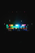 A vibrant stage setup featuring a large digital screen displaying a pattern of colorful blocks in shades of blue, yellow, red, and green. Silhouettes of musicians and equipment are visible in the foreground, creating a dynamic and energetic atmosphere.