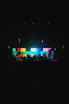 A vibrant stage setup featuring a large digital screen displaying a pattern of colorful blocks in shades of blue, yellow, red, and green. Silhouettes of musicians and equipment are visible in the foreground, creating a dynamic and energetic atmosphere.