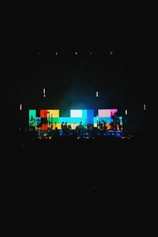 A vibrant stage setup featuring a large digital screen displaying a pattern of colorful blocks in shades of blue, yellow, red, and green. Silhouettes of musicians and equipment are visible in the foreground, creating a dynamic and energetic atmosphere.