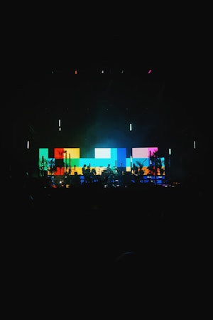 A vibrant stage setup featuring a large digital screen displaying a pattern of colorful blocks in shades of blue, yellow, red, and green. Silhouettes of musicians and equipment are visible in the foreground, creating a dynamic and energetic atmosphere.