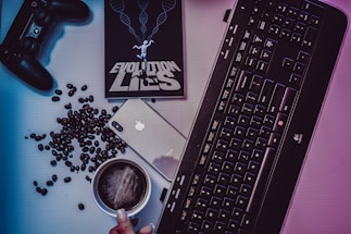 A desktop arrangement featuring a gaming controller, coffee beans scattered around, a smartphone with an Apple logo, a black keyboard, a cup of coffee, and a book titled 'EVOLUTION LIES' with a human figure and DNA helix imagery on the cover.