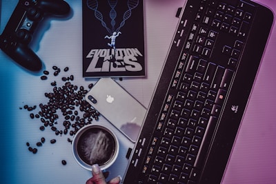 A desktop arrangement featuring a gaming controller, coffee beans scattered around, a smartphone with an Apple logo, a black keyboard, a cup of coffee, and a book titled 'EVOLUTION LIES' with a human figure and DNA helix imagery on the cover.