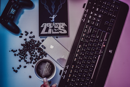 A desktop arrangement featuring a gaming controller, coffee beans scattered around, a smartphone with an Apple logo, a black keyboard, a cup of coffee, and a book titled 'EVOLUTION LIES' with a human figure and DNA helix imagery on the cover.