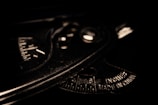 A close-up view of a metallic protractor or measuring tool with engraved measurements and text indicating it is made in China. The lighting is low, casting shadows and emphasizing the tool's industrial texture and markings.
