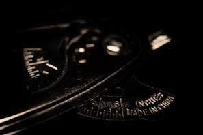 A close-up view of a metallic protractor or measuring tool with engraved measurements and text indicating it is made in China. The lighting is low, casting shadows and emphasizing the tool's industrial texture and markings.