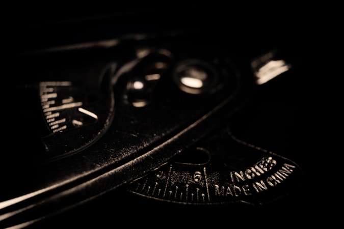 A close-up view of a metallic protractor or measuring tool with engraved measurements and text indicating it is made in China. The lighting is low, casting shadows and emphasizing the tool's industrial texture and markings.