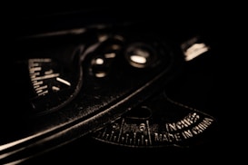A close-up view of a metallic protractor or measuring tool with engraved measurements and text indicating it is made in China. The lighting is low, casting shadows and emphasizing the tool's industrial texture and markings.