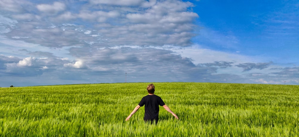 boy in black t-shirt standing on green grass field under white clouds and blue sky