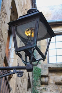 A vintage metal street lamp with a glass enclosure is mounted on an old stone wall. The lamp is slightly rusted, and a warm light glows from inside. The background shows a part of an old building with stone walls and a window with multiple panes.