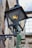 A vintage metal street lamp with a glass enclosure is mounted on an old stone wall. The lamp is slightly rusted, and a warm light glows from inside. The background shows a part of an old building with stone walls and a window with multiple panes.