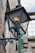 A vintage metal street lamp with a glass enclosure is mounted on an old stone wall. The lamp is slightly rusted, and a warm light glows from inside. The background shows a part of an old building with stone walls and a window with multiple panes.