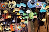 A variety of colorful, intricately designed mosaic lamps hang from the ceiling, showcasing vibrant patterns and a mix of shapes and sizes. The lamps are lit, casting a warm glow and creating a dynamic, artistic atmosphere.