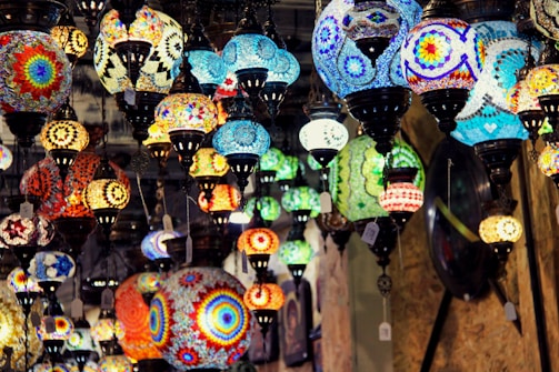 A variety of colorful, intricately designed mosaic lamps hang from the ceiling, showcasing vibrant patterns and a mix of shapes and sizes. The lamps are lit, casting a warm glow and creating a dynamic, artistic atmosphere.