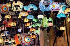 A variety of colorful, intricately designed mosaic lamps hang from the ceiling, showcasing vibrant patterns and a mix of shapes and sizes. The lamps are lit, casting a warm glow and creating a dynamic, artistic atmosphere.