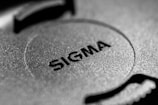 A close-up image of a textured surface featuring the word 'SIGMA' embossed in bold letters. The surface appears to be made of metal with a circular design element in the center.