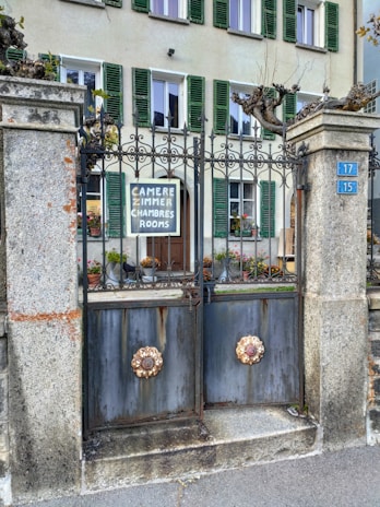 A wrought iron gate with ornate floral designs is flanked by stone pillars. Behind the gate, a building has windows with green shutters. A sign on the gate offers rooms in multiple languages. Potted flowers add a touch of color.