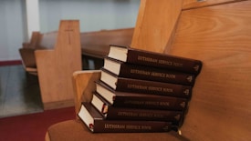 A stack of Lutheran service books is placed neatly on a wooden church pew, with more pews visible in the background. The setting suggests a quiet, serene church interior with an empty, peaceful atmosphere.