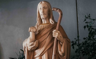 A wooden statue of a robed figure with long hair holding a lamb in one arm and a shepherd's crook in the other. The figure has a serene expression and is surrounded by leafy plants.