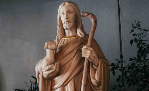 A wooden statue of a robed figure with long hair holding a lamb in one arm and a shepherd's crook in the other. The figure has a serene expression and is surrounded by leafy plants.