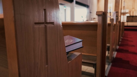 A wooden pew with a carved cross on the side panel is seen in a church. On the edge of the pew rests a closed book with a cross imprinted on its cover. The interior of the church is softly lit, featuring a red carpet aisle leading towards an altar in the background.