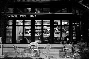 A black and white photograph of a wine bar with a glass front, showing the interior where people are working behind the counter. The shelves are lined with numerous glasses and bottles. Outside, there are a few metal chairs and tables on a small patio, surrounded by potted plants.