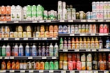 A grocery store shelf filled with a wide variety of colorful beverages in cans and bottles. The products include coconut water, carbonated soft drinks, teas, and fruit-flavored drinks. The packaging features vibrant colors and different languages.
