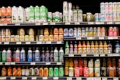 A grocery store shelf filled with a wide variety of colorful beverages in cans and bottles. The products include coconut water, carbonated soft drinks, teas, and fruit-flavored drinks. The packaging features vibrant colors and different languages.