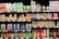 A grocery store shelf filled with a wide variety of colorful beverages in cans and bottles. The products include coconut water, carbonated soft drinks, teas, and fruit-flavored drinks. The packaging features vibrant colors and different languages.