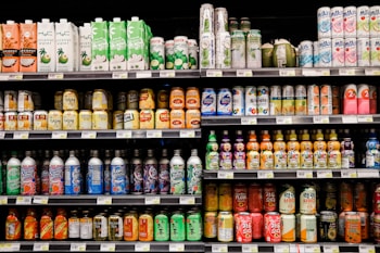 A grocery store shelf filled with a wide variety of colorful beverages in cans and bottles. The products include coconut water, carbonated soft drinks, teas, and fruit-flavored drinks. The packaging features vibrant colors and different languages.